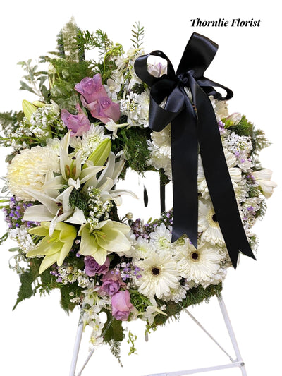 Funeral wreath with white lilies, purple roses, and white gerberas with a black ribbon.